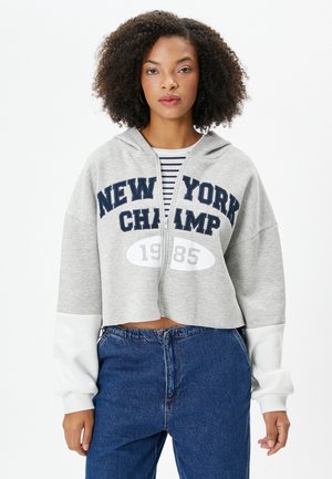Geknipt grijze hoodie met marineblauwe tekst "NEW YORK CHAMP" en het jaar "1985." Witte mouwen, ritssluiting aan de voorkant, casual pasvorm, zachte stof.