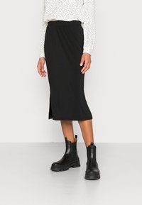 Jupe midi noire avec fente sur le côté, en matériau doux, associée à des bottines noires épaisses à bout carré avec une semelle épaisse et des poignées.