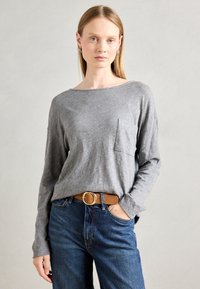 Kvinne med rett blondt hår iført en grå langermet topp med brystlomme, blå jeans og et brunt belte med gullspenne, stående innendørs.
