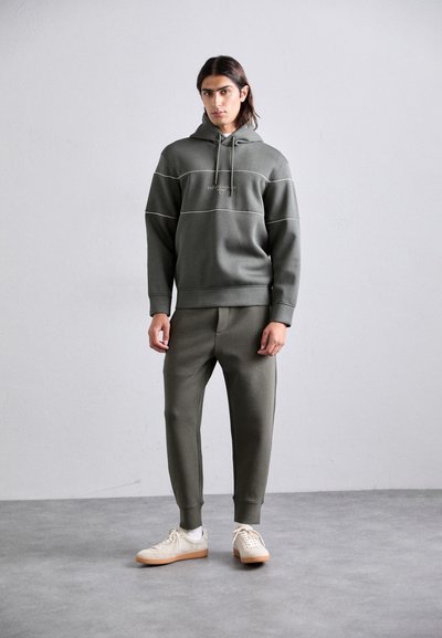 Hanorac de culoare verde măslin cu șnur și cusături accentuate albe, asortat cu pantaloni jogger potriviți; material neted și croială lejeră, purtat cu adidași crem.