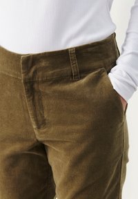 Olijfgroene corduroy broek met een brede tailleband, zijzakken en een subtiele ribbelstructuur. Combinatie met een strak wit shirt met lange mouwen.