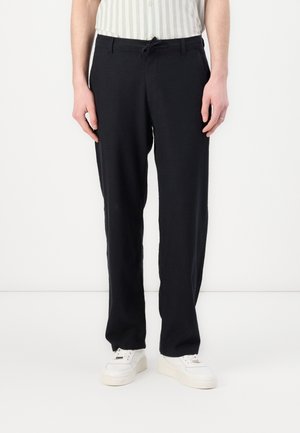 Man i svarta raka byxor med knytmidja och vita sneakers, stående mot en vit bakgrund.