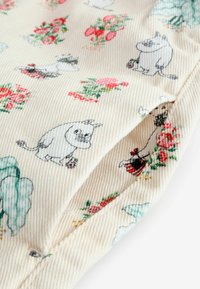 Krém háttérrel rendelkező pamut anyag, amely színes Moomin karaktereket és virágmintákat tartalmaz. Bordázott textúrájú zöld és piros részletekkel.