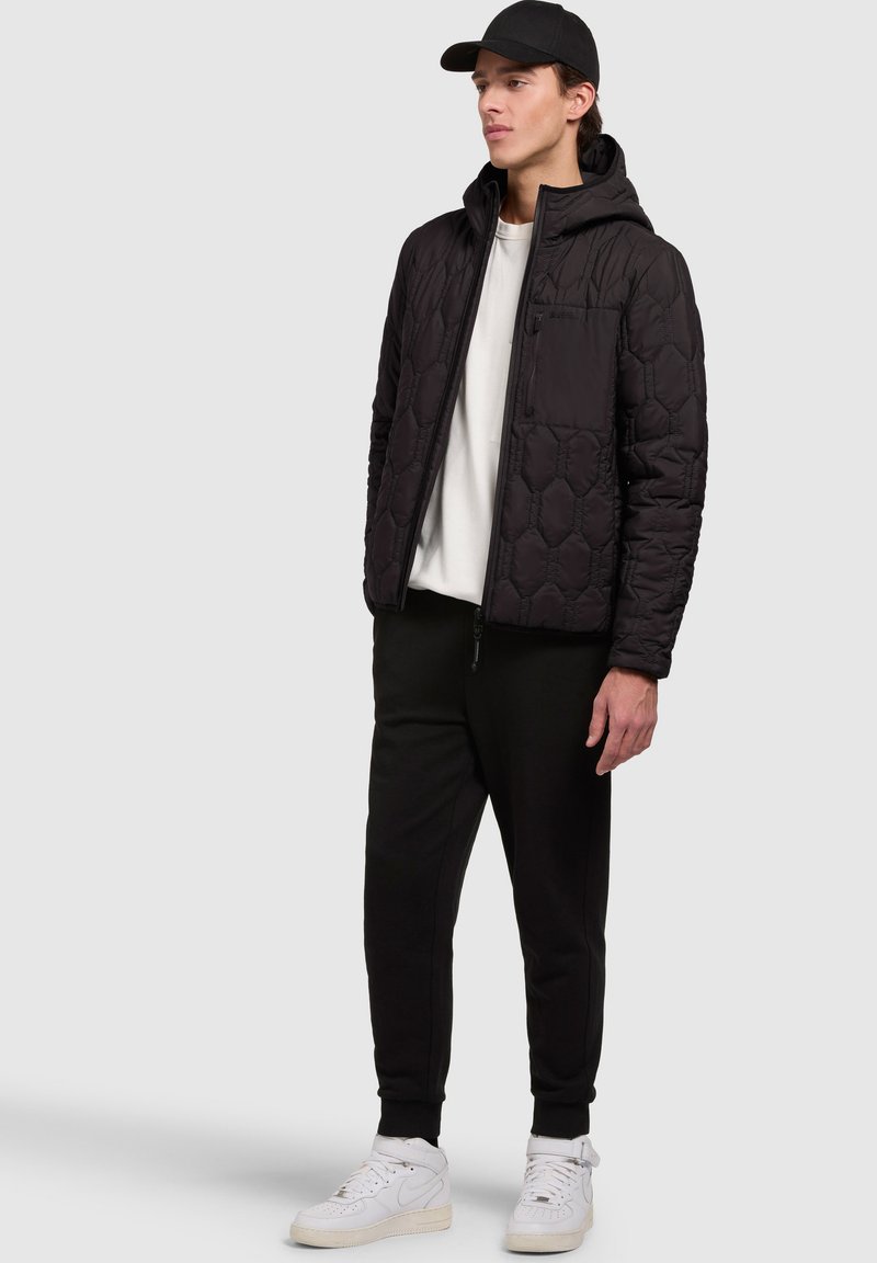 Veste noire matelassée avec capuche, dotée d'une fermeture éclair et d'un motif hexagonal, portée sur une chemise blanche, associée à un pantalon noir et à des baskets blanches.
