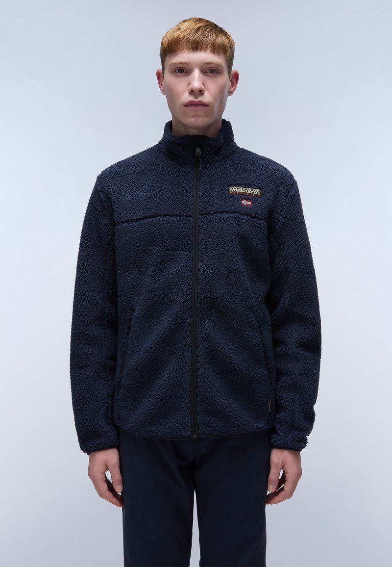 Marineblaue Fleecejacke mit hohem Kragen, durchgehendem Reißverschluss und strukturierter Oberfläche. Fällt auf durch ein Logo-Patch und seitliche Taschen.