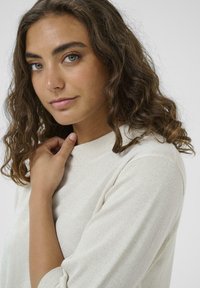 Svetlo béžový sveter s jemným leskom, s okrúhlym výstrihom a trojštvrťovými rukávmi, nosený osobou s kučeravými vlasmi.