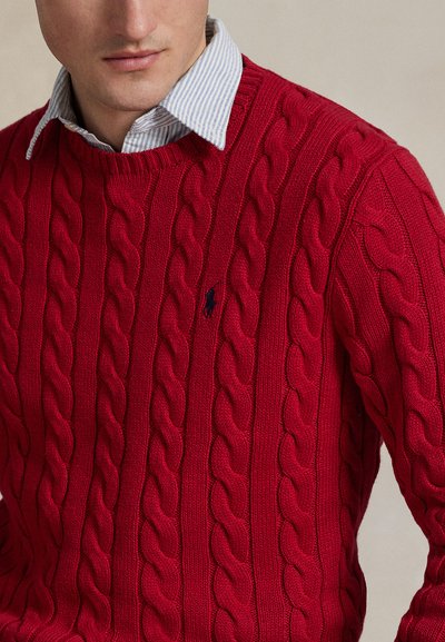 Uomo che indossa un maglione rosso a trecce sopra una camicia a righe azzurra chiaro con colletto, ritagliato alle spalle e alla bocca
