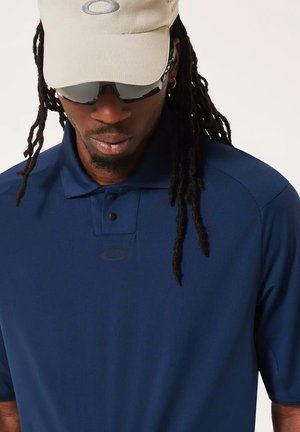 Man with long dreadlocks wearing navy blue polo shirt, beige cap, and black wraparound sunglasses, looking downward.