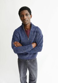 Blauer Pullover mit hohem Kragen und Reißverschluss, mit strukturiertem Strickmuster. Kombiniert mit grauen Jeans steht das Modell mit verschränkten Armen.