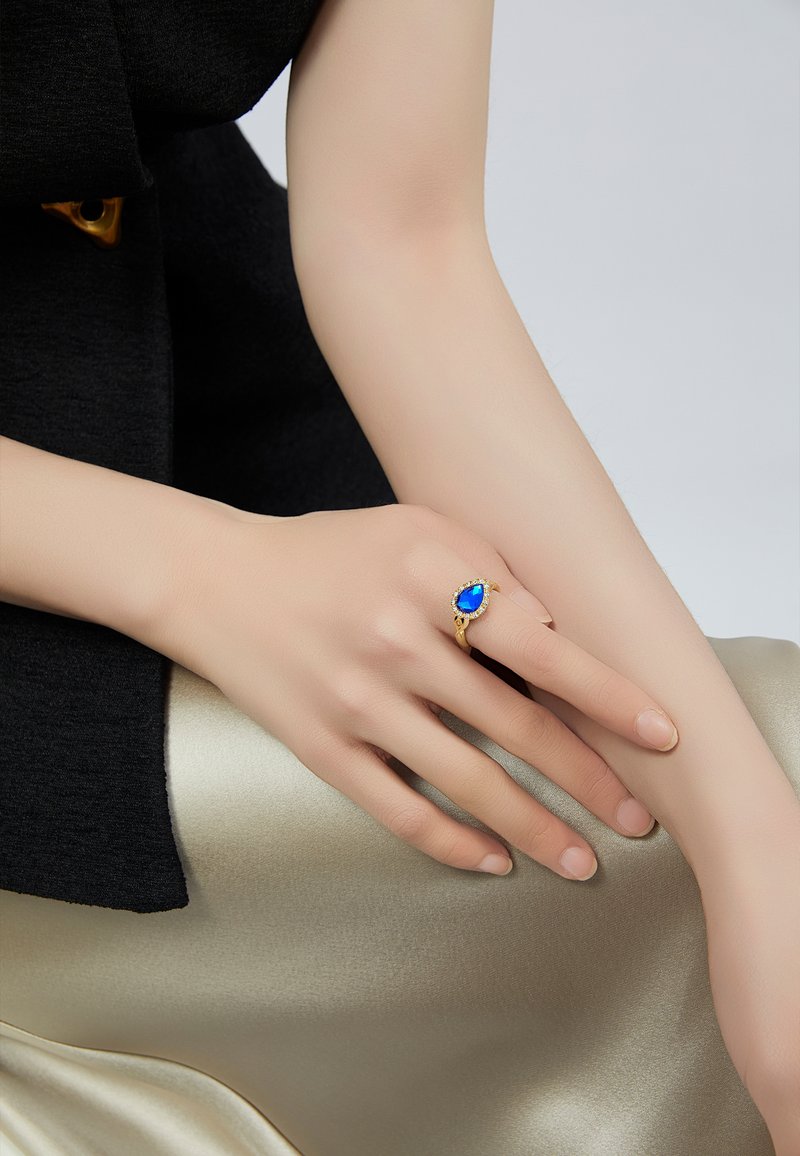 Anello d'oro con una pietra blu a forma di pera, circondata da piccoli gemme trasparenti. La mano poggia su una superficie dorata morbida e lucida. Indumento nero testurizzato sullo sfondo.