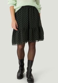 Jupe noire à volants avec un motif floral vert subtil, confectionnée en tissu léger. Associée à des bottines noires et des collants transparents.