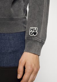 Sudadera gris con puños acanalados, que presenta un logo bordado de "HUG" con un corazón. Mano visible, descansando sobre una tela de mezclilla.