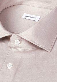 Chemise beige clair à boutons avec col pointu et boutons blancs, montrant l'étiquette de la marque "seidensticker" à l'intérieur du col.