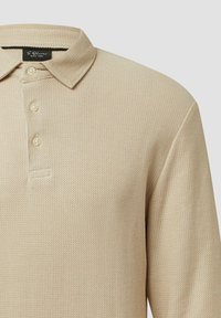 Beżowa koszulka polo z teksturowanym materiałem dzianinowym, z klapką na trzy guziki i klasycznym kołnierzykiem. Dyskretne detale logo w pobliżu kołnierza.