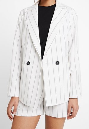 Witte blazer en korte broek met dunne verticale grijze strepen, zwarte knopen op de blazer, gedragen over een zwarte top, afgebeeld van halverwege de dij tot aan de nek.