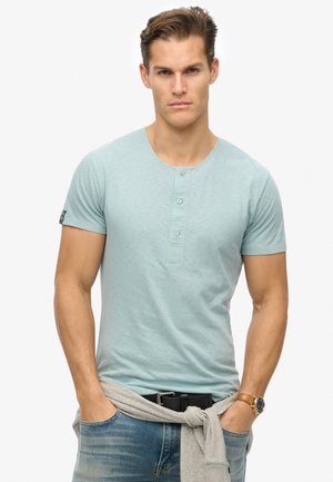 Light blue short-sleeve henley shirt with three button placket and soft texture; worn with gray sweater tied around the waist.