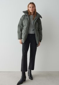 Giacca a vento verde scuro con colletto in finta pelliccia, abbinata a un maglione grigio chiaro, jeans neri a vita alta e stivali neri alla caviglia.