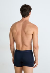 Homem em pé de costas, sem camisa, vestindo boxers azul-escuro contra um fundo claro simples.