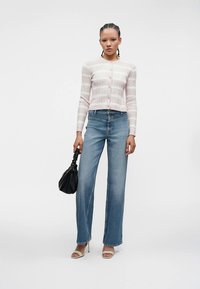 Γυναίκα φορώντας ανοιχτό ροζ ριγέ ζακέτα, ψηλόμεσες φαρδιές μπλε τζιν, λευκά ανοιχτόκαρδα ψηλά τακούνια, κρατώντας μαύρη τσάντα με ρυτιδώσεις.