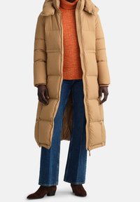 Lange tan pufferjas met capuchon, met een gewatteerd ontwerp. Draag het over een feloranje geribd sweater en wijde denim jeans. Bruine enkellaarsjes.