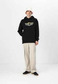 Black hoodie with graphic print on the front, featuring a kangaroo pocket. Paired with beige trousers and black sneakers. White bucket hat.