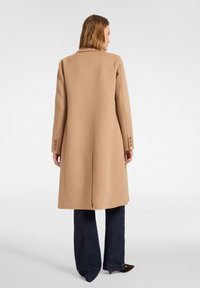 Mujer con un abrigo largo color camel con puños abotonados, jeans oscuros de pierna ancha y zapatos marrones puntiagudos, de pie de espaldas sobre un fondo blanco.