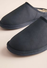 Paire de chaussons bleu foncé à enfiler avec dessus texturé et doublure intérieure douce, posés sur une surface de couleur claire.