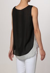Blusa nera senza maniche con retro trasparente e orlo curvo, indossata sopra una base grigia chiara. Abbinata a pantaloni bianchi.