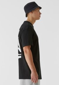 Black cotton t-shirt with short sleeves, featuring a large white graphic on the back. Model also wears a navy bucket hat.