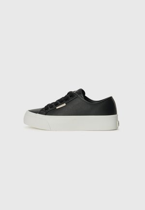 Lavtop sort sneakers med hvid tyk sål, sorte snørebånd og lille metal logo mærke på siden, vist på en ensfarvet lys baggrund.