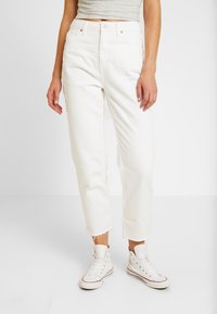 Žena v bílých džínech s vysokým pasem a raw lemy, v kombinaci s bílými vysokými teniskami a béžovým zkráceným topem na jednobarevném pozadí.