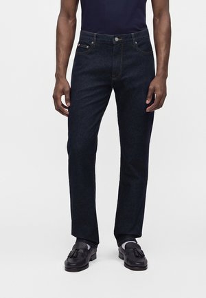 Mand iført mørkeblå straight-leg jeans, sorte loafers med duske og en marineblå skjorte, stående mod en ensfarvet lys baggrund.