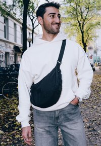 Borsa a tracolla nera in tessuto con forma curvata, cinturino regolabile e superficie texture, indossata sopra un hoodie bianco e jeans grigi.