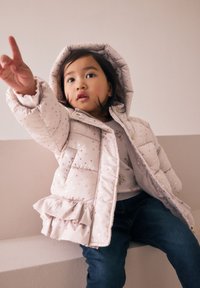 Veste matelassée rose clair avec motif d'étoiles dorées, capuche et ourlet à volants, portée par-dessus une chemise à fleurs et un jean foncé.