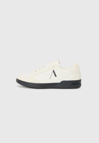 Zapatilla deportiva blanca con un upper texturizado, suela negra y un acento geométrico en el costado. Los cordones con un diseño plano y redondeado completan el aspecto.