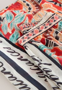 Tissu coloré avec des motifs floraux et abstraits complexes en rouge, sarcelle, noir et blanc, avec une partie plissée et un texte en écriture noire.