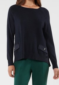 Pull à manches longues bleu marine avec un col rond, un ourlet côtelé et deux poches avant ornées de boutons décoratifs. Texture douce et coupe décontractée.