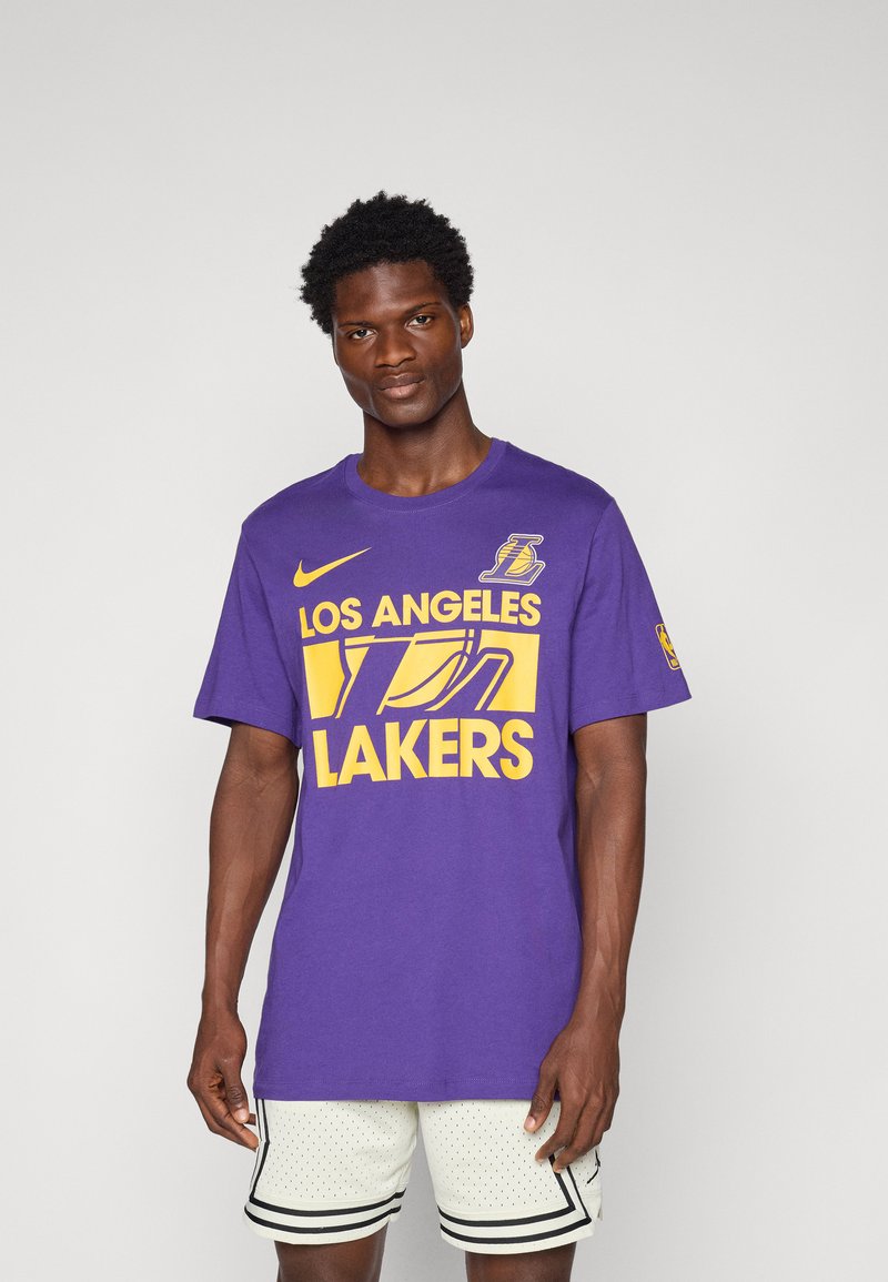 Purpura kokvilnas T-krekls ar uzrakstu "LOS ANGELES LAKERS" dzeltenā krāsā, ar apaļu kaklu un īsām piedurknēm. Uz priekšpuses ir Nikes logo.