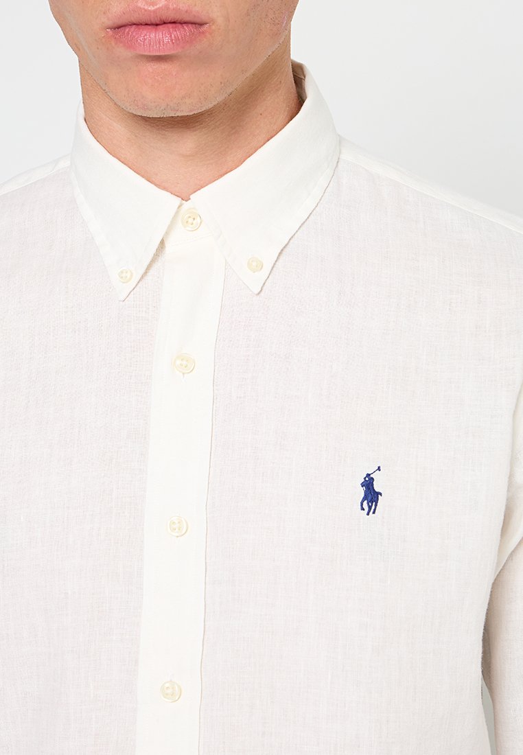 Chemise en lin blanche à boutons avec un col classique, présentant une texture subtile et un logo brodé bleu sur la poitrine gauche.