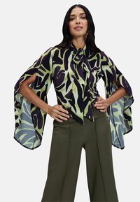 La blusa multicolore presenta un motivo astratto verde e nero, maniche larghe e un dettaglio al collo con nodo, abbinata a pantaloni verde oliva.