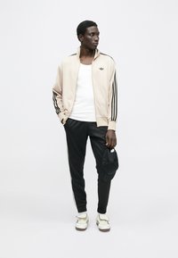 Bež sportska jakna na patentni zatvarač s crnim prugama, bijela majica bez rukava, crne trenerke i kremasti tenisice, drži crnu kapu u jednoj ruci.