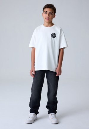 Teenager trägt ein weißes übergroßes T-Shirt mit rundem schwarzem Logo, schwarze Jeans und weiße Sneakers, steht vor einem schlichten hellen Hintergrund.