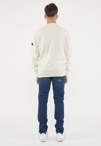 Maglione lavorato a maglia beige chiaro con scollo rotondo e maniche lunghe; abbinato a jeans in denim blu scuro con un motivo decorativo a punto.