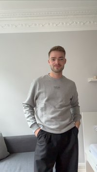 Siva sweatshirt s krožno ovratnikom, z črnim potiskom na prsih, v kombinaciji s črnimi hlačami. Gladka tekstura, sproščen kroj.
