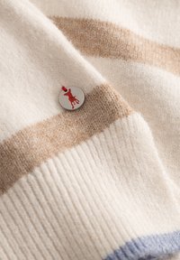 Kremowa i beżowa wełniana tkanina w paski o prążkowanej teksturze, z okrągłą srebrną metką z sylwetką czerwonego psa oraz czerwoną wstążką.