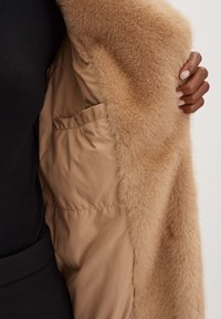Cappotto beige foderato in pelliccia sintetica con un tessuto interno liscio. Presenta una tasca laterale e una texture morbida e soffice lungo il bordo e la foderatura.