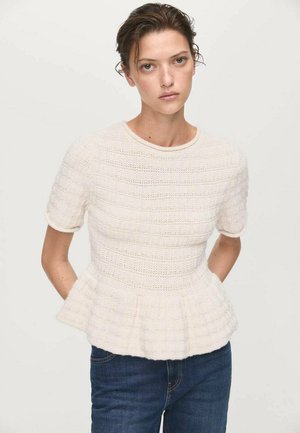 Donna con capelli castani che indossa un maglione bianco a peplo con maniche corte tessurate e jeans blu, su uno sfondo chiaro e uniforme.