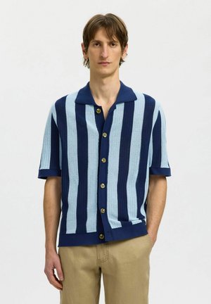 Mann iført kortermet, knappet strikkeskjorte med vertikale lyse og mørkeblå striper, kombinert med beige bukser, stående foran en ensfarget bakgrunn.