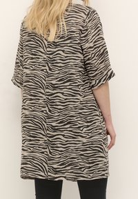 Beige tunika dengan garis zebra hitam, lengan pendek, dan potongan lurus. Kainnya tampak lembut dengan sedikit tekstur.