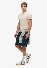 Beżowa koszulka z okrągłym dekoltem, granatowe szorty, jasnoszary sweter związany w talii, białe adidasy z czarnymi akcentami i białe skarpetki. Niezobowiązujący strój.